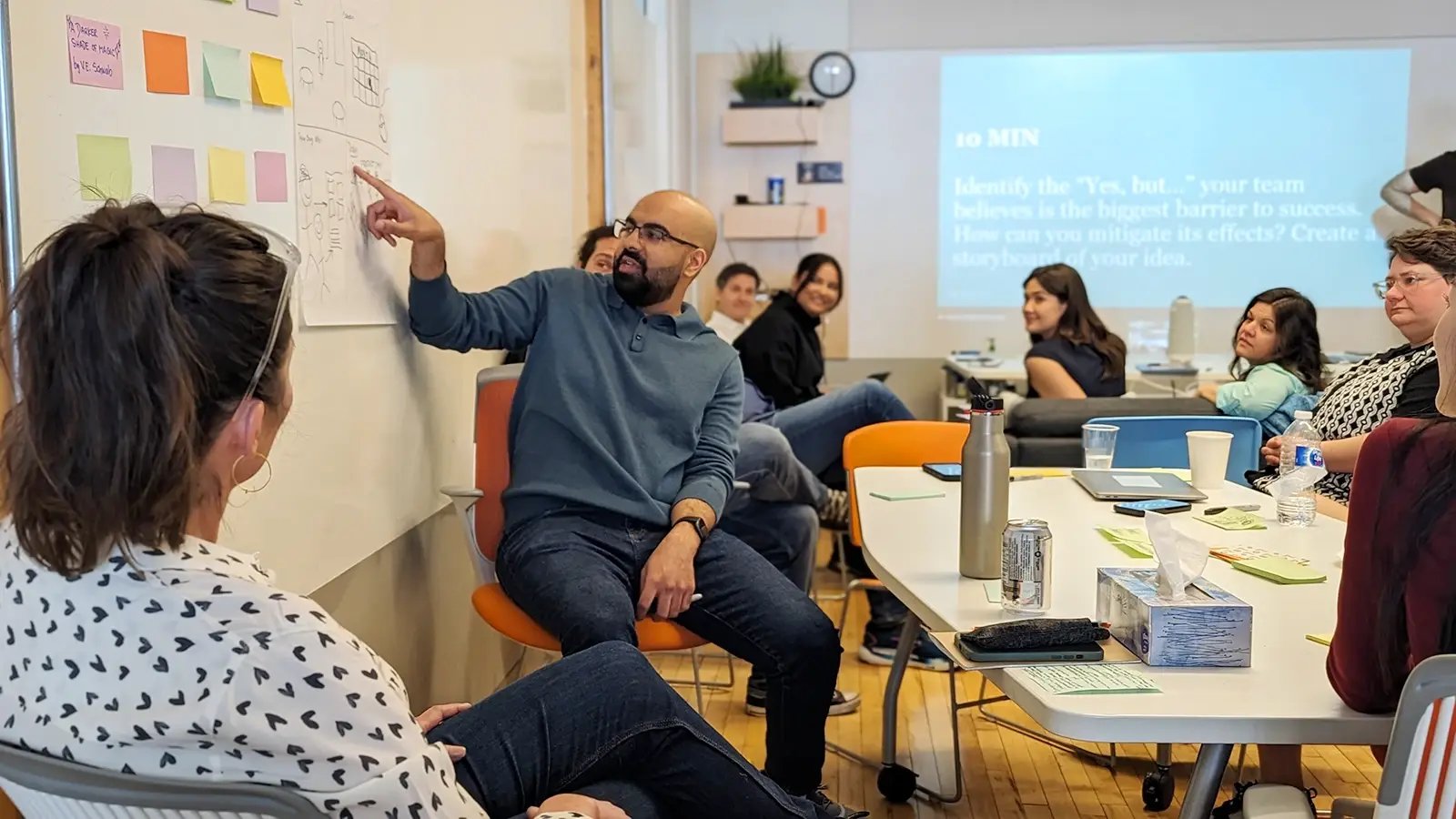 A team member seated in a collaborative workspace points at a storyboard on a whiteboard, engaging with colleagues who are also seated around tables.