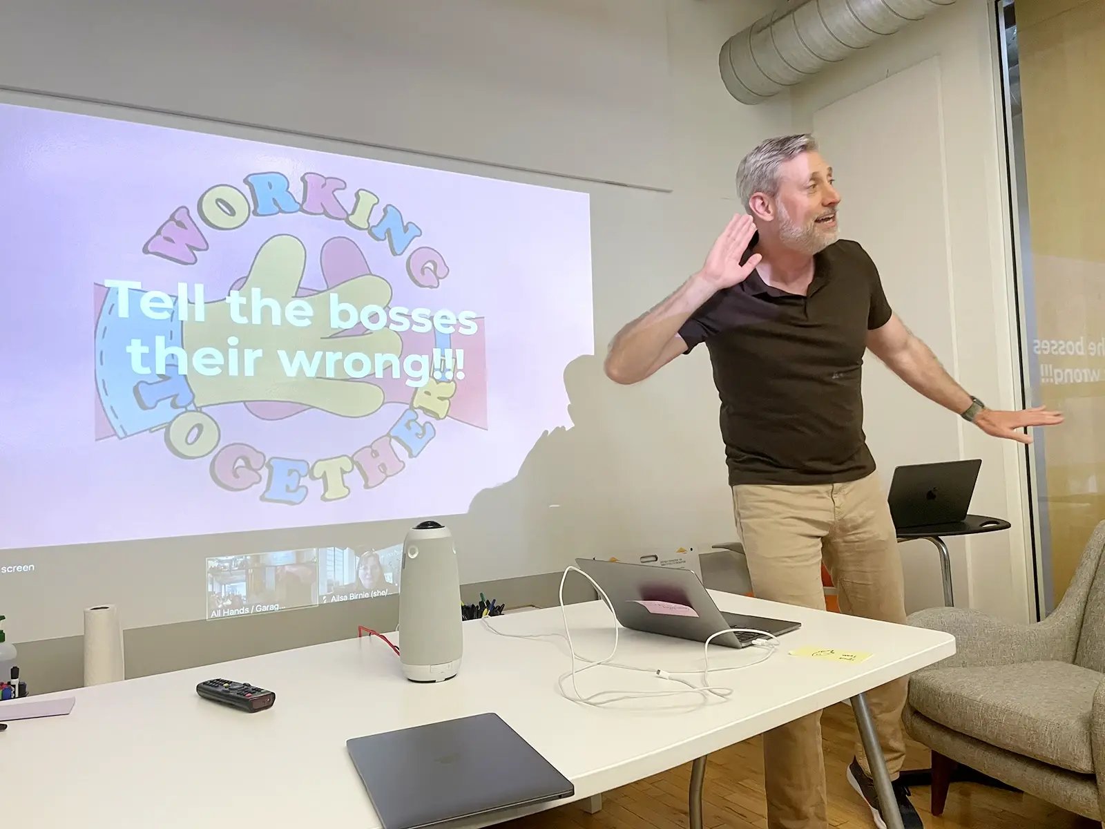 ExperiencePoint co-founder Greg Warman stands before a projected presentation screen, energetically gesturing with one hand raised to his ear. The screen displays the text "Tell the bosses their wrong!!!" over a colorful "WORKING TOGETHER" graphic.